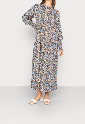 Frau trägt ein langärmliges, knöchellanges Blumenkleid mit blauen, gelben und weißen Blumen und weißen Sandalen mit Absatz, steht auf grauem Boden.