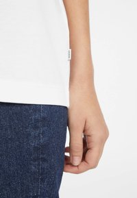Maglietta bianca a maniche corte con etichetta laterale, abbinata a jeans in denim scuri. Il tessuto appare liscio e leggero, evidenziando un design casual.