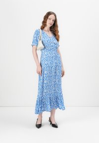 Robe maxi bleue à fleurs avec des manches courtes à volants, taille cintrée et ourlet évasé. Associée à des escarpins noirs à bout pointu et un sac à main blanc.