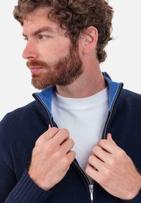 Pull à fermeture éclair bleu marine avec col intérieur bleu, tissu texturé et poignets côtelés. Le modèle tient la fermeture éclair devant un fond blanc.