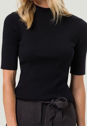 Femme portant un pull noir côtelé à manches courtes et un pantalon gris taille haute avec une ceinture en tissu nouée à la taille.
