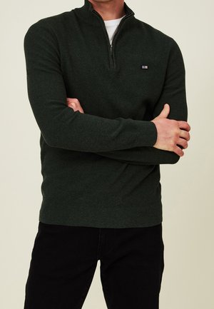 Homme portant un pull vert foncé à fermeture éclair sur le haut avec un petit logo sur la poitrine, un sous-vêtement blanc et un pantalon noir, les bras croisés devant un fond beige.