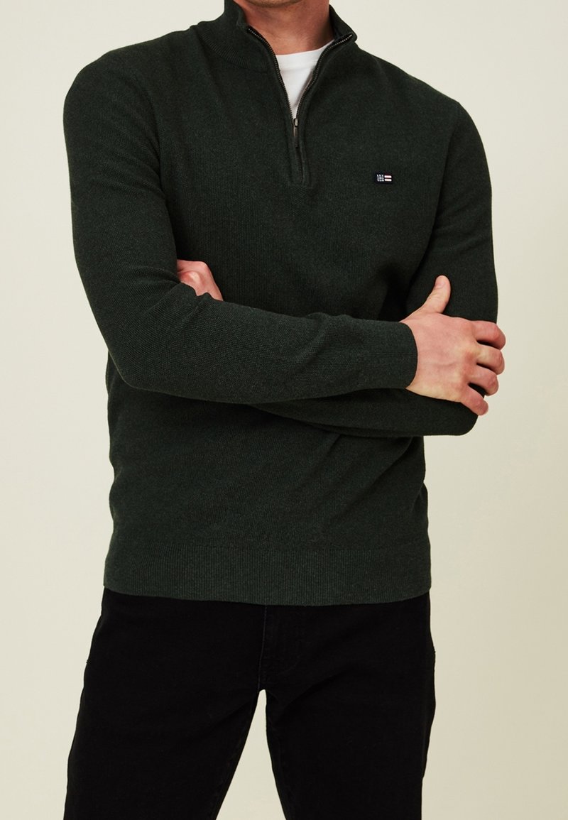 Homme portant un pull vert foncé à fermeture éclair sur le haut avec un petit logo sur la poitrine, un sous-vêtement blanc et un pantalon noir, les bras croisés devant un fond beige.