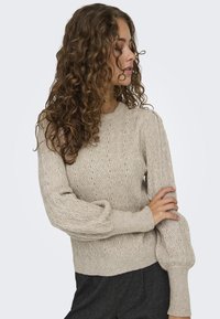 Pull en tricot beige clair avec un motif en vagues, doté de manches longues légèrement bouffantes et de poignets en maille côtelée. Tissu texturé avec des détails subtils.