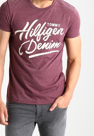 Hombre con una camiseta color burdeos de Tommy Hilfiger Denim y vaqueros grises, con una mano en el bolsillo y la otra descansando a su lado.