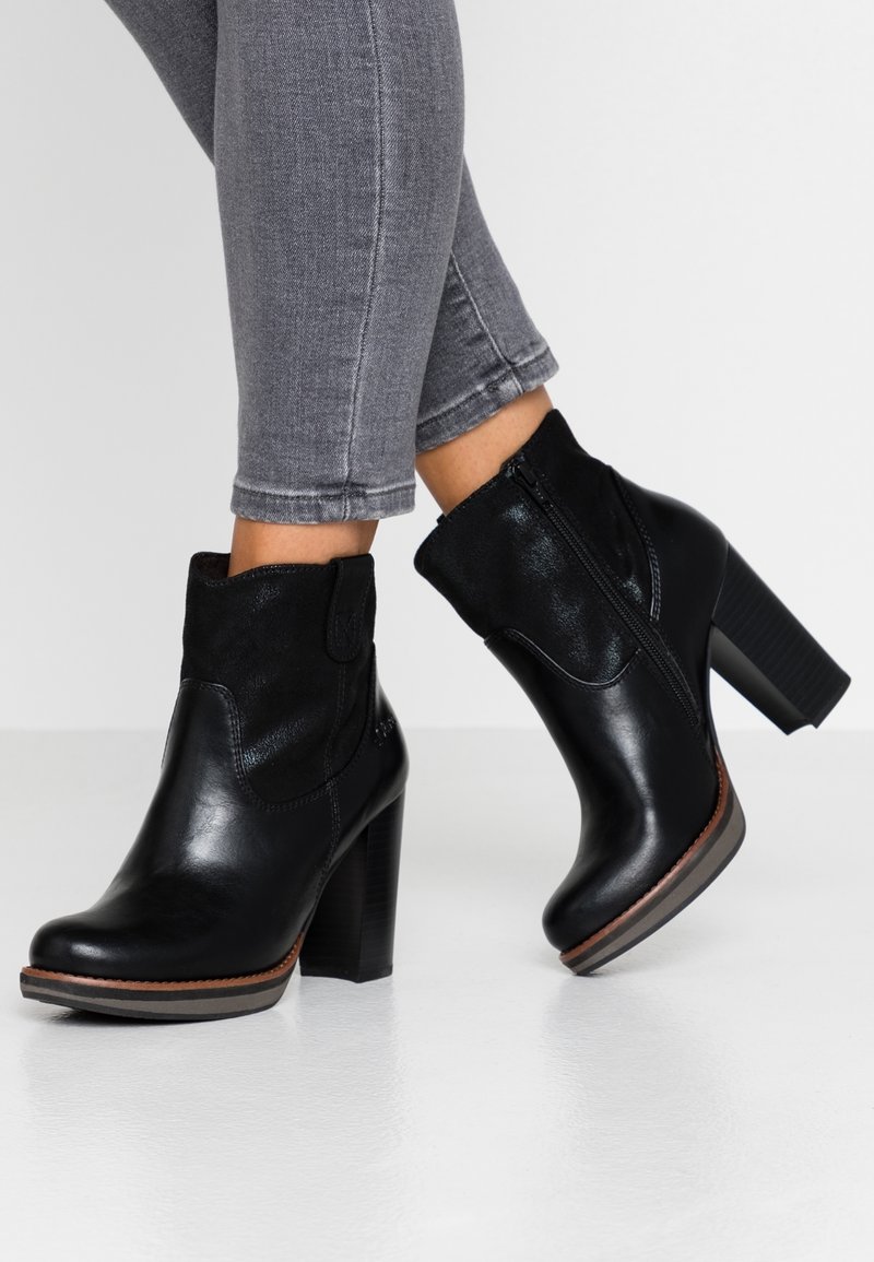 Personne portant des bottines en cuir noir à talons carrés et fermetures éclair sur les côtés, associées à un jean gris ajusté sur un fond uni.