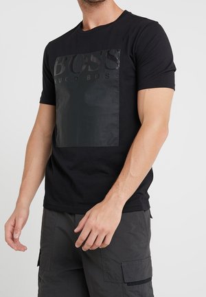 Mann trägt ein schwarzes Hugo Boss T-Shirt mit einem glänzenden schwarzen Logo-Patch und dunkelgrauen Cargoshorts vor einfarbigem Hintergrund.