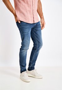 Jeans de mezclilla azul con un corte slim, que presentan un ligero desgaste y costuras en contraste, combinados con zapatillas blancas y una camisa de cuadros rosas.
