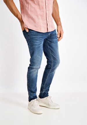 Jean bleu en denim avec une coupe slim, présentant un léger délavage et des coutures contrastantes, associé à des baskets blanches et une chemise à carreaux rose.
