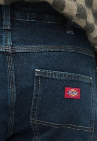 Σκούρο μπλε τζιν με τσέπη στην κάτω πλάτη που φέρει κόκκινο λογότυπο "Dickies" και αντίθετες κίτρινες ραφές.