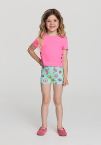 Camicetta rosa a maniche corte con dettagli di taglio laterale, abbinata a pantaloni corti verde menta con motivi colorati di ciambelle e fenicotteri. Scarpe rosa.