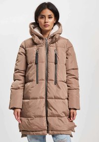 Ung kvinne iført en lang, beige polstret jakke med stor hette, to vertikale glidelåslommer, og slitte lyseblå jeans.