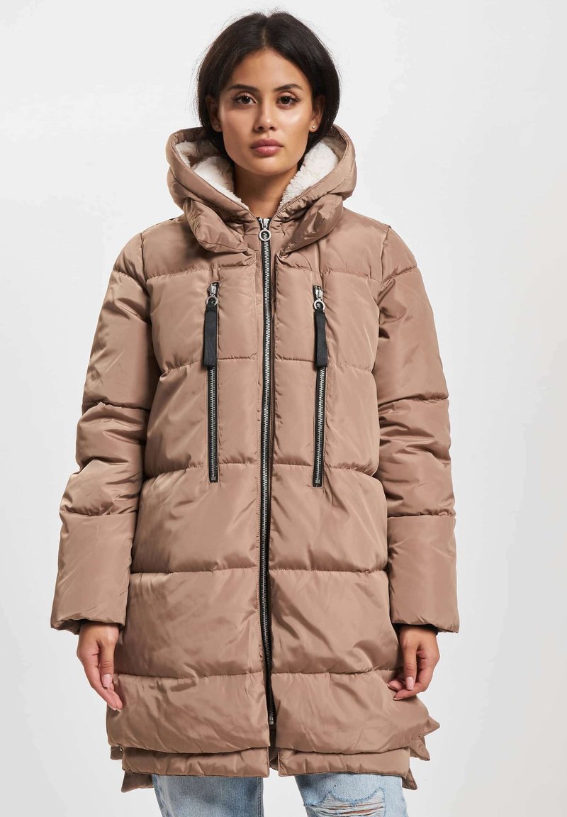 Ung kvinne iført en lang, beige polstret jakke med stor hette, to vertikale glidelåslommer, og slitte lyseblå jeans.