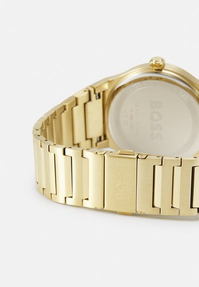 Montre-bracelet de couleur or avec un bracelet en métal à maillons rectangulaires. Le dos présente un verre transparent et la marque "BOSS".