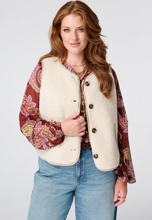Vrouw met lang kastanjebruin haar die een crèmekleurig fleecevest met knopen draagt over een rode paisley-blouse en een blauwe spijkerbroek, zacht glimlachend.