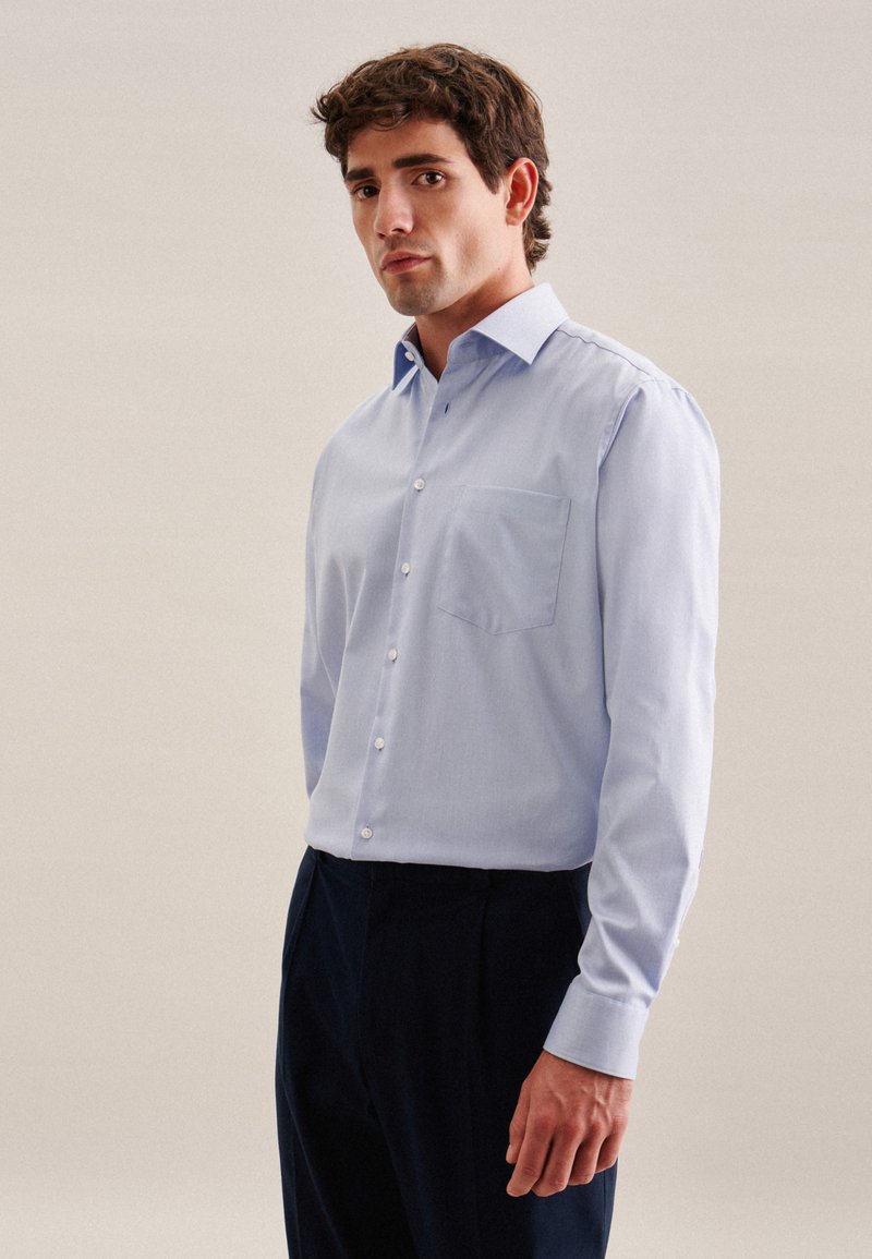 Chemise à manches longues bleu clair avec un col à boutons, dotée d'une poche poitrine et d'un tissu lisse et texturé. Associée à un pantalon sombre.
