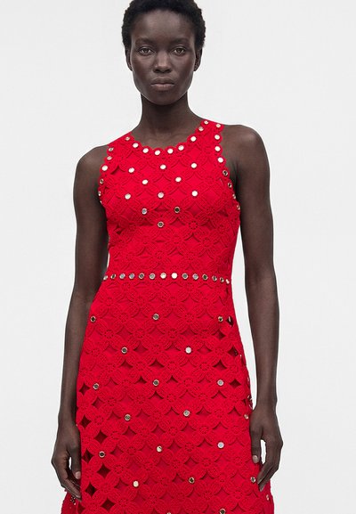 Femme portant une robe rouge en dentelle sans manches avec un motif floral et de petites embellissements circulaires réfléchissants à l'avant, debout devant un fond uni.