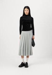 Fekete, hosszú ujjú, textúrált bársonytopper, amely egy pliszírozott szürke midi szoknyával van párosítva. A modellen fekete slip-on cipő és egy fekete táska van.