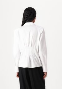 Blusa blanca con corte ajustado, que presenta una espalda plisada, acentos cosidos y mangas largas. Tejido suave, diseño de cuello clásico.