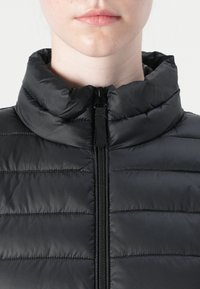 Svart polstret vest med høy krage, med sentral glidelås og quiltet tekstur. Materialet ser glinsende og lett ut.