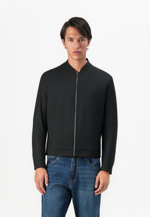 Man wearing a black zipped bomber jacket and blue jeans standing against a white background.
