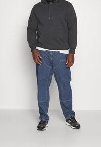 Sweat à capuche gris foncé avec une poche avant, superposé sur une chemise blanche ; associé à un jean bleu avec des coutures avant courbées et des baskets noires.