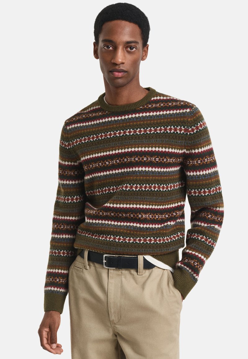 Maglione lavorato a maglia di colore verde scuro con strisce e fantasie orizzontali multicolori. Presenta un collo rotondo e polsini a costine. Abbinato a pantaloni kaki.