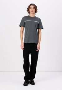 Grå t-skjorte med korte ermer og hvit logo stripe; kombinert med svarte jeans og sorte sko, stående mot en nøytral bakgrunn.