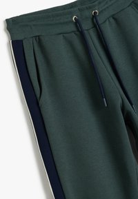 Donkergroene joggers met een geribde tailleband, marineblauwe zijkanten en witte accenten. Beschikt over een trekkoord en een zachte stofstructuur.