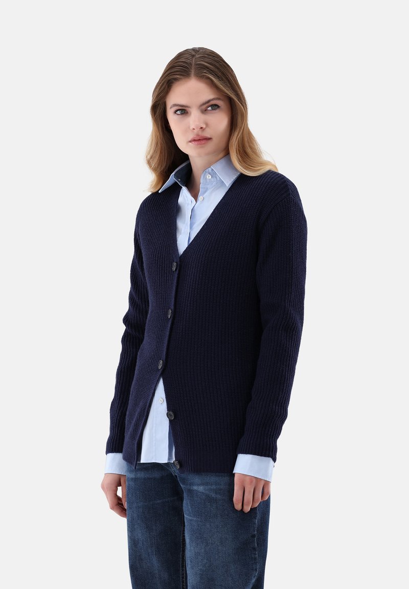 Marineblaue, gerippte Strickjacke mit V-Ausschnitt, fünf Knöpfen an der Vorderseite, strukturiertem Stoff, getragen über einem hellblauen Hemd mit Kragen.