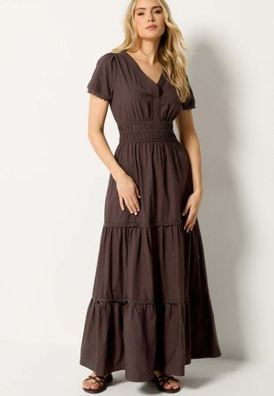 Vrouw draagt een donkerbruine, gelaagde maxi-jurk met korte mouwen, V-hals, geplooide taille en sandalen, staand tegen een effen achtergrond.