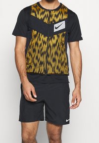 Sportliches Herrenoutfit mit einer Gilett mit Reißverschluss, gelb-schwarzem Muster, schwarzen kurzen Ärmeln und einem Nike-Logo auf der Brust.