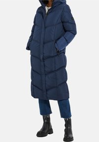 Lange marineblauwe pufferjas met capuchon, met een gequilt ontwerp, zijzakken en een rechte snit. Gecombineerd met cropped denim en laarzen.