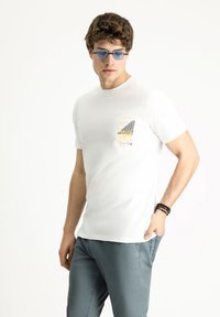 Camiseta blanca de algodón con un estampado gráfico en el pecho, combinada con pantalones azul claro. El modelo lleva gafas de sol azules en forma ovalada y una pulsera de cuentas.