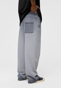 Grijze oversized sweatpants met een textuurafwerking, elastische tailleband en een patchzak op de achterkant. Gemaakt met zwarte sneakers.