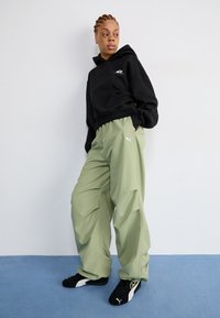 Black hooded sweatshirt with front pocket and logo, paired with loose, light green pants featuring side pleats, and black sneakers with white accents.