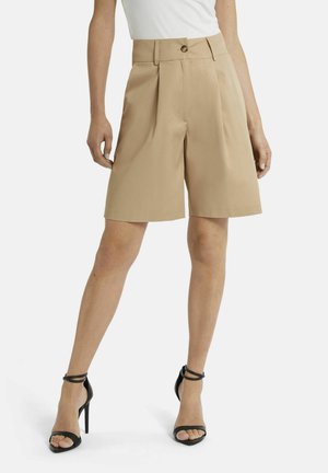 Modell trägt beige, hoch taillierte maßgeschneiderte Shorts mit vorderen Falten, kombiniert mit schwarzen High Heels mit Knöchelriemen und einem weißen, eingesteckten Oberteil.