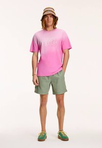 Rosa gradient t-shirt med texten "TRANQUILO", kombinerad med olivgröna shorts och en flerfärgad bucket-hatt, tillsammans med skor i teal och gult.
