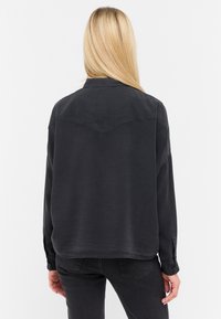 Camisa de mezclilla negra con un diseño de yugo puntiagudo, puños abotonados y un ajuste relajado. La tela tiene una textura suave con un acabado ligeramente desgastado.