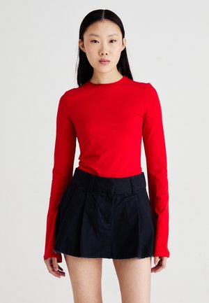 Crvena majica dugih rukava s okruglim izrezom, u kombinaciji s crnom plisiranom mini suknjom. Elegantan dizajn s glatkim teksturama i prilagođenim siluetom.