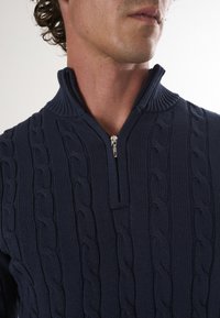Sweater tricoté noir marine avec un col montant, fermeture éclair à l'avant et un motif en câble texturé. Tissu lisse et épais avec des accents côtelés.