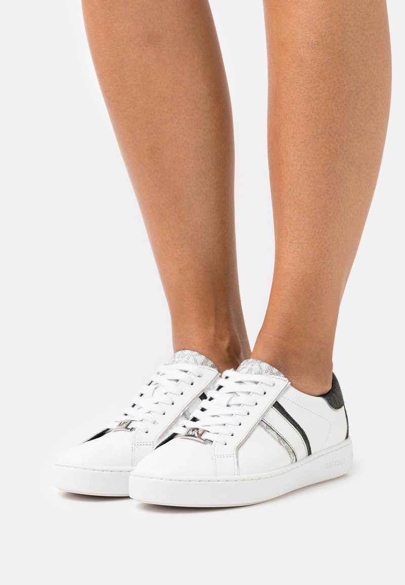 Białe sneakersy z srebrnymi i czarnymi akcentami, charakteryzujące się gładką skórzaną fakturą, okrągłym noskiem i płaską gumową podeszwą. Sznurowadła z przodu.