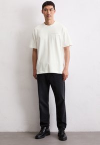 Biała koszulka bawełniana z małym nadrukiem, zestawiona z czarnymi dżinsami i czarnymi butami eleganckimi, na jednolitym tle.