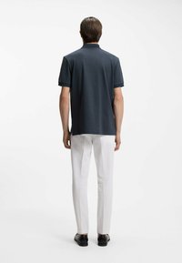 Granatowa koszulka polo z krótkimi rękawami, prążkowana tekstura, boczne rozcięcia, w połączeniu z białymi spodniami i czarnymi skórzanymi butami. Widok od tyłu.