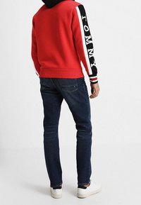 Man in een rode hoodie met zwart-witte mouwen waarop de tekst "TOMMY" staat, donkere spijkerbroek en witte sneakers, staande met zijn rug naar de kijker gericht.