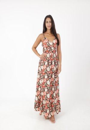 Donna in piedi con una mano sul fianco, indossa un lungo vestito floreale con spalline sottili, su uno sfondo bianco semplice.