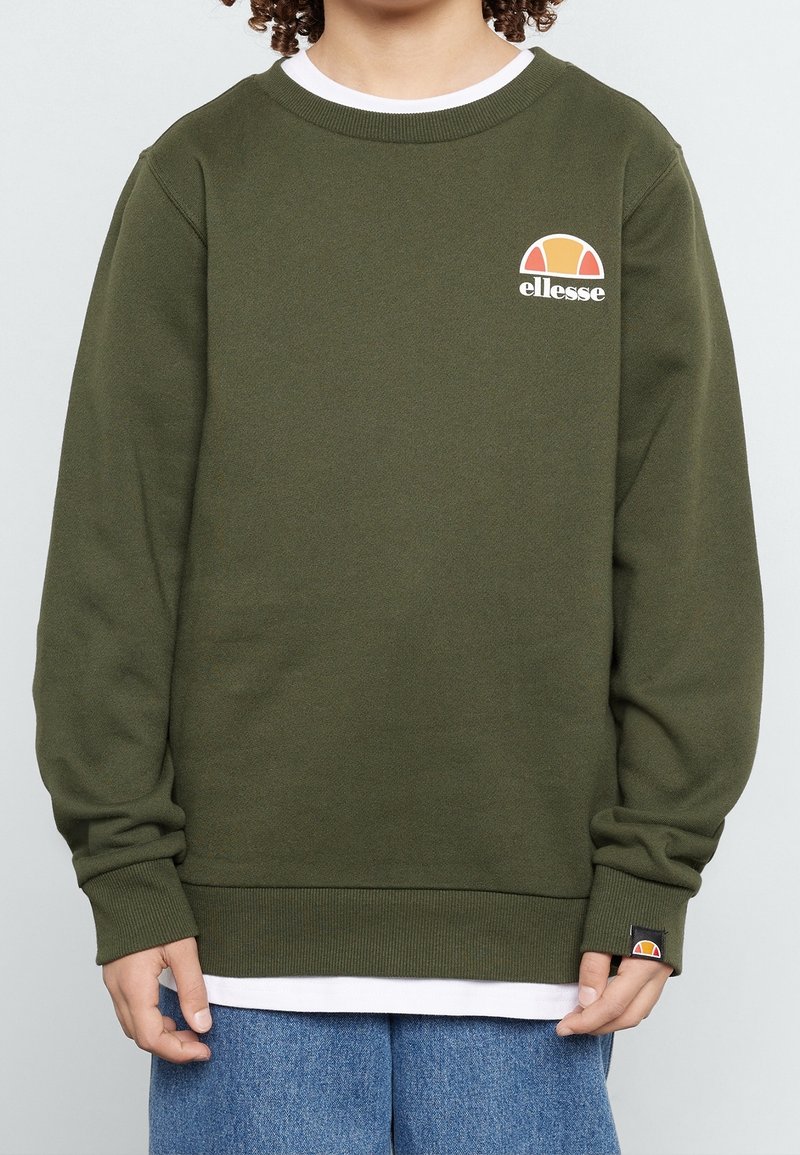 Enfant portant un sweat-shirt ellesse vert olive avec un col blanc et un jean bleu, debout devant un fond clair uni.