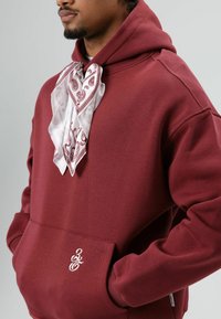 Bordeauxrode hoodie met een voorzak, met een witte sjaal met paisley-patroon gestrikt om de hals en een klein geborduurd logo op de zak.