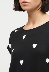 Γυναίκα που φοράει μαύρο T-shirt με μικρά λευκά σχέδια καρδιάς, εμφανιζόμενη από τον λαιμό μέχρι το επάνω μέρος του στήθους.