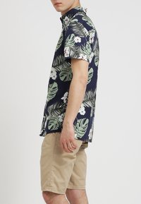 Košile s krátkým rukávem na knoflíky v námořnické modři s potiskem zelených tropických listů a bílých květin, v kombinaci s béžovými kraťasy. Bavlněná látka, volný střih.